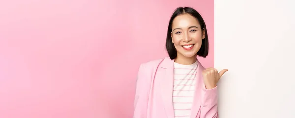 정장을 입은 외국인 사업가가 웃는 모습, 하얀 빈 벽을 손가락으로 가리키는 기업 여성의 모습, 정보나 광고를 든 보드, 핑크 색 배경 위에 서 있는 모습 — 스톡 사진