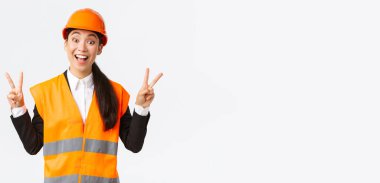Mutlu gülümseyen Asyalı kadın mühendis, güvenlik giysileri ve kasklı mimar, barış kawaii işareti gösteriyor ve neşeli görünüyor, inşaat ve inşaat ihalesini kazanan şirket, beyaz arka plan