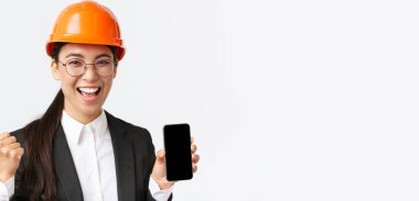 Başarılı bir Asyalı kadın mühendis, güvenlik kaskı ve giysisi içinde mimar, akıllı telefon ekranı, yumruk pompası ve evet diye bağırma, başarıya karşı zafer, beyaz arka plan
