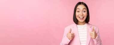 Asyalı iş kadını portresi tatmin olmuş gülümsüyor, başparmaklarını gösteriyor, övüyor ve onaylıyor, pembe arka planda takım elbise içinde duruyor.