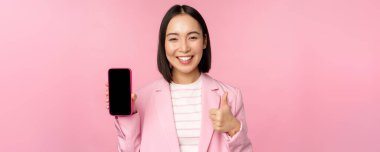 Tatmin olmuş gülümseyen Asyalı iş kadını cep telefonu uygulaması öneriyor, akıllı telefondan web sitesi şirketi, ekranı ve başparmakları gösteriyor, pembe arka plan