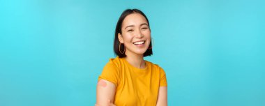 Covid-19 'dan aşı kampanyası. Mutlu ve sağlıklı Asyalı kız Coronavirüs aşısından sonra gülüyor, omzunda bandaj, sarı tişört, mavi arka plan