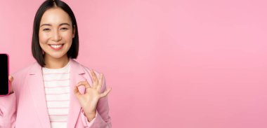 Tatmin olmuş gülümseyen Asyalı iş kadını cep telefonu uygulaması öneriyor, akıllı telefondan web sitesi şirketi ekran ve tamam işareti gösteriyor, pembe arka plan