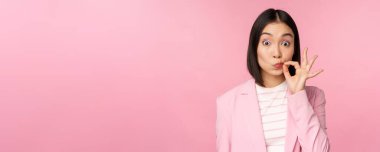Asyalı kurumsal bir kadının portresi, ağzı mühürlü, dudakları kapalı anahtar hareketi, söz ver sır tut, pembe arka planda duran takım elbiseli.