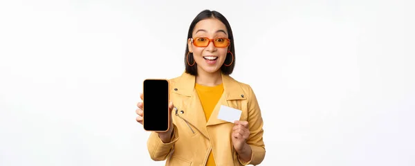 Zakupy online i koncepcja ludzi. Stylowa azjatycka kobieta pokazująca ekran telefonu komórkowego i kartę kredytową, aplikację na smartfona, stojąca na białym tle — Zdjęcie stockowe