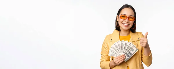 Snygg leende asiatisk flicka som håller pengar kontanter, visar dollar och firar, står över vit bakgrund — Stockfoto