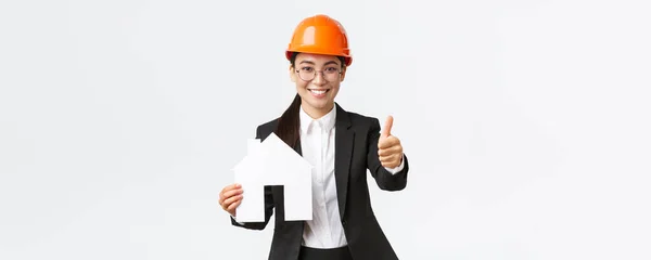 Retrato de jovem asiático engenheiro de construção feminino, arquiteto em capacete de segurança e terno de negócios segurando casa maket e mostrar polegares para cima, promovendo o serviço da empresa de construção de casas — Fotografia de Stock