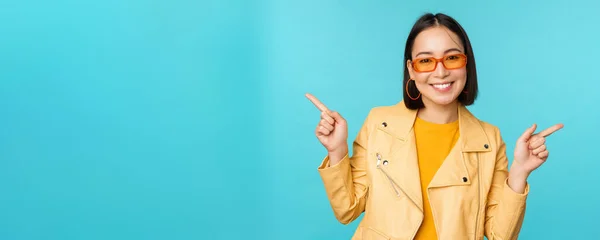 Menina chinesa elegante feliz em óculos de sol, aponta os dedos lateralmente, convida para check-out, ver desconto na loja, mostra esquerda e direita, fica sobre fundo azul — Fotografia de Stock