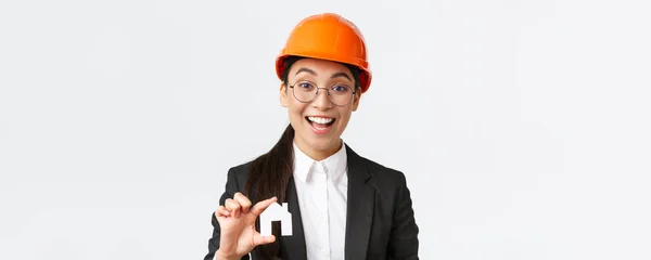 Primer plano del corredor profesional en casco de seguridad y traje de negocios que vende casas. Alegre arquitecto mostrando minuatura de casa y sonriendo, trabajando sobre nuevo diseño o construcción, fondo blanco —  Fotos de Stock