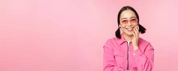Detailní portrét stylové asijské ženy ve slunečních brýlích, usmívající se, tvářící se polichoceně, smějící se koketně, stojící nad růžovým pozadím — Stock fotografie