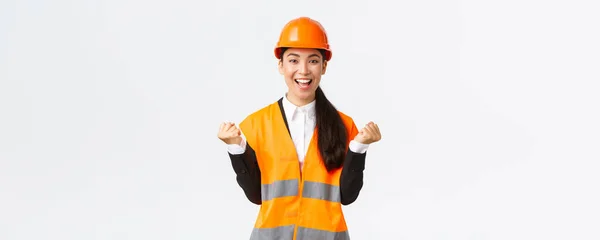 Succès gagnant asiatique femme gestionnaire de la construction, ingénieur à la vue du bâtiment portant un casque de sécurité, levant les mains dans la joie, pompe poing de la réalisation et la victoire, fond blanc — Photo