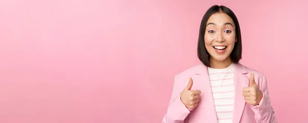 Πορτρέτο της ασιάτισσας επιχειρηματία χαμογελά ικανοποιημένη, δείχνει τους αντίχειρες επάνω, επαίνους, όπως και να εγκρίνει, στέκεται στο κοστούμι πάνω από ροζ φόντο — Φωτογραφία Αρχείου