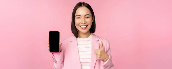 Soddisfatto sorridente donna d'affari asiatica che raccomanda app per telefoni cellulari, società di siti web su smartphone, mostrando schermo e pollici in su, sfondo rosa — Foto Stock