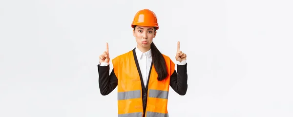 Byggnads-, bygg- och industrikoncept. Missnöjd sorgsen asiatisk kvinnlig arkitekt klagar, bär skyddshjälm och reflekterande kläder, sura upprörda som pekar finger upp — Stockfoto