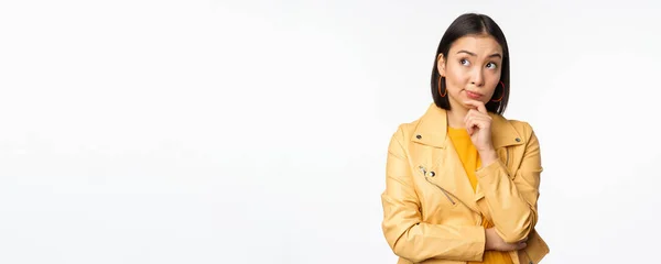 黄色のジャケットでスタイリッシュな韓国語の女性の肖像画、思慮深い笑顔、思考とロゴや広告を見上げ、白い背景の上に立って — ストック写真