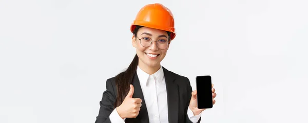 Primo piano di ingegnere femminile sorridente professionista, responsabile della costruzione in tuta da lavoro e casco di sicurezza, che mostra lo schermo dello smartphone e i pollici in alto in approvazione, sfondo bianco — Foto Stock