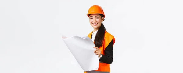 Приємна усміхнена жінка азіатська менеджерка будівництва після креслень під час будівництва будинку, дивлячись задоволена камерою, вивчає архітекторський план, стоїть білий фон в шоломі безпеки — стокове фото
