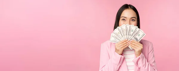 Feliz ásia senhora no terno segurando dinheiro, dólares com satisfeito rosto expressão, pé sobre rosa fundo — Fotografia de Stock