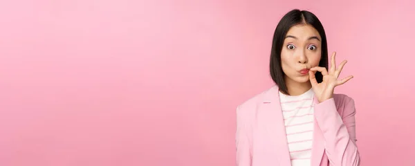 Портрет азійської корпоративної жінки з печаткою рота, закриті губи за ключовим жестом, обіцяйте тримати в секреті, стоячи над рожевим фоном в костюмі — стокове фото