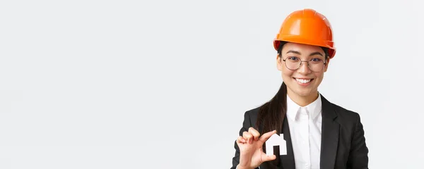 행복 한 아시아인 여성의 근접 사진, 헬멧과 비지니스 슈트를 입고 미니어처와 미소를 지닌 부동산 중개업자, 건물 보수 작업을 하는 건축가, 백인 배경 — 스톡 사진