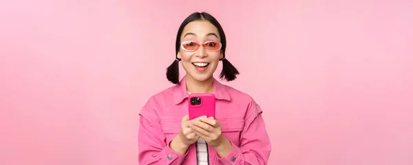 Ritratto di ragazza coreana con eleganti occhiali da sole che reggono il telefono cellulare, utilizzando l'app per smartphone, in piedi su sfondo rosa — Foto Stock