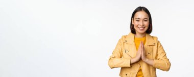 Mutlu Koreli kadın, umut dolu görünüyor, yardım istiyor, yalvarıyor, namaste jesti ile duruyor ve gülümsüyor, beyaz arka planda duruyor.
