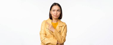 Asyalı kız şüpheci görünüyor, şüpheli bir şekilde solu işaret ediyor, şüpheli ve hoşnutsuz bakıyor, beyaz arka planda duruyor.