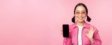 Hevesli genç Asyalı kadın tamam, tamam işareti, gülümseme memnun, cep telefonu ekranı, akıllı telefon uygulaması, pembe arka planda duruyor.