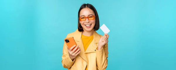 Compras en línea. Elegante mujer asiática joven en gafas de sol, mostrando la tarjeta de crédito y el uso de teléfono inteligente, pagando en Internet, haciendo la compra, de pie sobre fondo azul — Foto de Stock