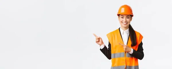 Bâtiment, construction et concept industriel. Souriant asiatique femme architecte dans casque de sécurité, vêtements réfléchissants pointant du doigt le coin supérieur gauche, montrant le projet sur le lieu de travail, fond blanc — Photo