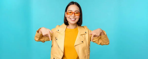 Asyalı şık bir kızın portresi, güneş gözlüğü takıyor, gülümsüyor ve parmağıyla işaret ediyor, reklamını yapıyor, mavi arka planda duruyor. — Stok fotoğraf