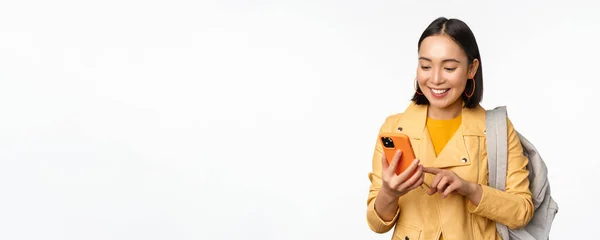Imagen de chica viajera feliz, turista con mochila mirando el teléfono inteligente, utilizando la aplicación de ruta del teléfono móvil, de pie sobre fondo blanco —  Fotos de Stock