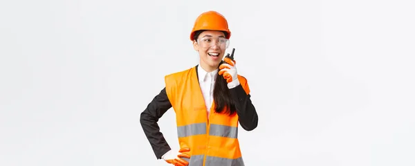 Confident pleased asian female engineer in safety helmet and uniform talking with chief architect using walkie-talkie. Satisfied construction technician contact team using radio phone — Stock Photo, Image