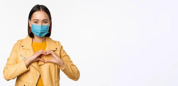 Covid-19 och människor koncept. Ung asiatisk kvinna i medicinsk ansiktsmask, visar hjärtat gest och ler med omsorg, vit bakgrund — Stockfoto