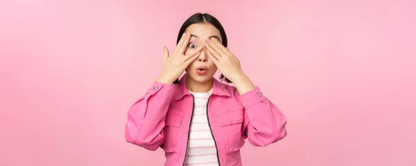 Portré ázsiai lány kukucskál izgalommal ujjakon keresztül, takarja szem, látva meglepetés, álló felett rózsaszín háttér — Stock Fotó