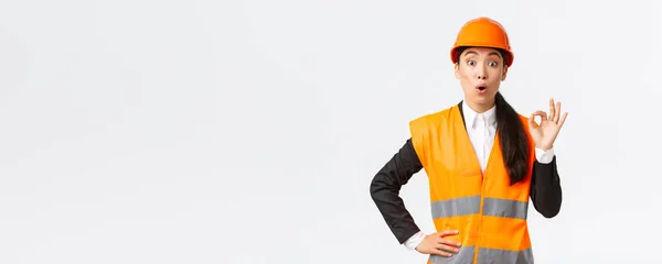 Imponerad och förvånad asiatisk kvinnlig byggchef nöjd med fantastisk kvalitet, står i skyddshjälm och reflekterande jacka, visar okej gest och säga wow, vit bakgrund — Stockfoto
