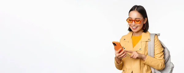 Elegante giovane donna asiatica turista, viaggiatore con zaino e smartphone sorridente alla macchina fotografica, in posa su sfondo bianco — Foto Stock