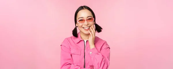 Close up retrato de mulher asiática elegante em óculos de sol, sorrindo, olhando lisonjeado, rindo coquete, de pé sobre fundo rosa — Fotografia de Stock