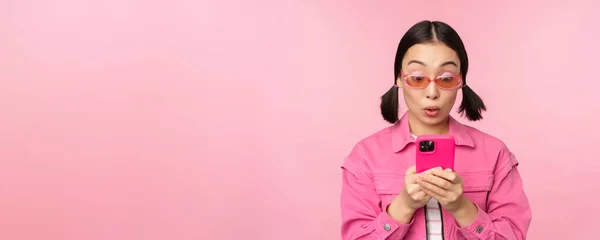 Retrato de chica asiática en gafas de sol usando smartphone. Mujer mirando el teléfono móvil, la navegación en la aplicación, de pie sobre el fondo rosa — Foto de Stock