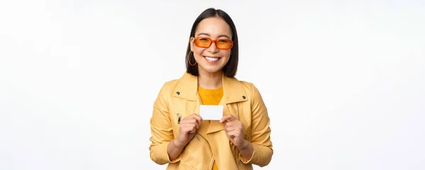 Portrét krásné moderní asijské dívky ve slunečních brýlích, šťastný úsměv, ukazující kreditní kartu, stojící nad bílým pozadím — Stock fotografie
