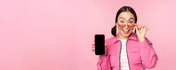 Ritratto di ragazza asiatica che mostra lo schermo del telefono cellulare, reagendo sorpreso, in piedi su sfondo rosa — Foto Stock