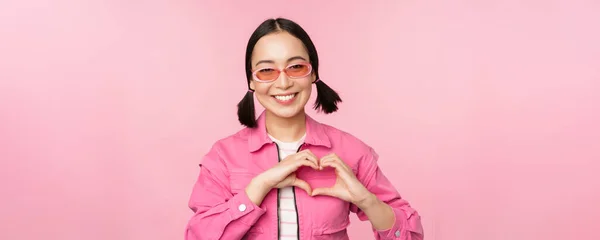 Preciosa modelo femenina coreana en gafas de sol de moda, muestra el corazón, signo de cuidado, te amo gesto, se para sobre fondo rosa — Foto de Stock