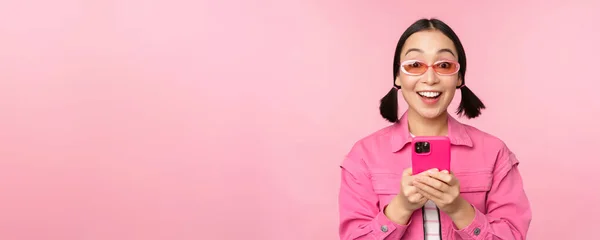 Retrato de chica coreana en gafas de sol elegantes que sostienen el teléfono móvil, utilizando la aplicación del teléfono inteligente, de pie sobre el fondo rosa —  Fotos de Stock