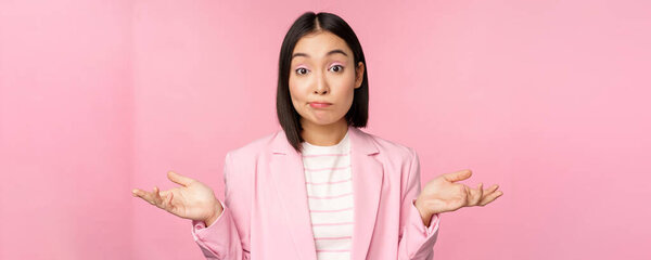 Portrait of confused asian businesswoman shrugging shoulders, looking clueless and puzzled, dont know, cant say, standing over pink background in office suit