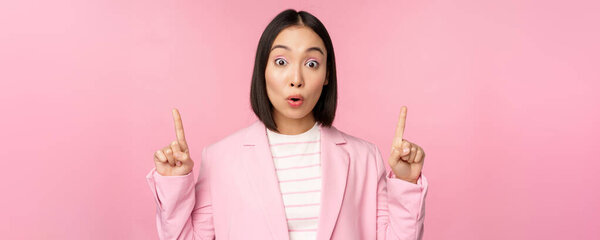 Portrait of asian businesswoman, corporate worker in suit, looks surprised by advertisement, points finger up, showing banner or logo on top, stands over pink background