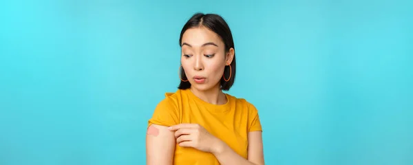 Campagna vaccinale da Covid-19. Giovane bella, sana donna asiatica mostrando spalla con bandaid, concetto di vaccinazione, in piedi su sfondo blu — Foto Stock