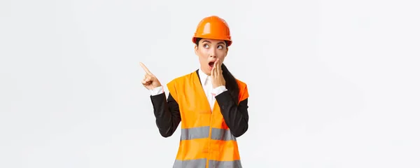身穿安全帽和反光夹克的亚洲女工程师震惊而印象深刻，气喘吁吁地指着左上角，惊奇地发现了一些有趣的东西 — 图库照片