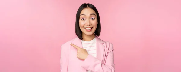 Portrét asijské podnikatelky, prodavačka v obleku ukazující prstem vlevo, ukazující reklamu na banner, usmívající se a vypadající profesionální, růžové pozadí — Stock fotografie