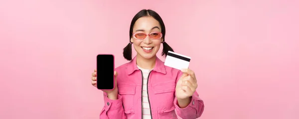 Зображення усміхненої корейської жінки, що показує кредитну картку і екран мобільного телефону, інтерфейс смартфонів, оплата онлайн, безконтактні покупки, стоячи над рожевим фоном. — стокове фото