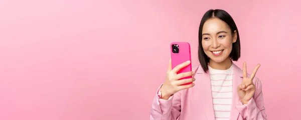 Şık Asyalı iş kadını, takım elbiseli kız akıllı telefondan selfie çekiyor, cep telefonu uygulaması ile görüntülü konuşma, pembe stüdyo geçmişine karşı poz veriyor. — Stok fotoğraf